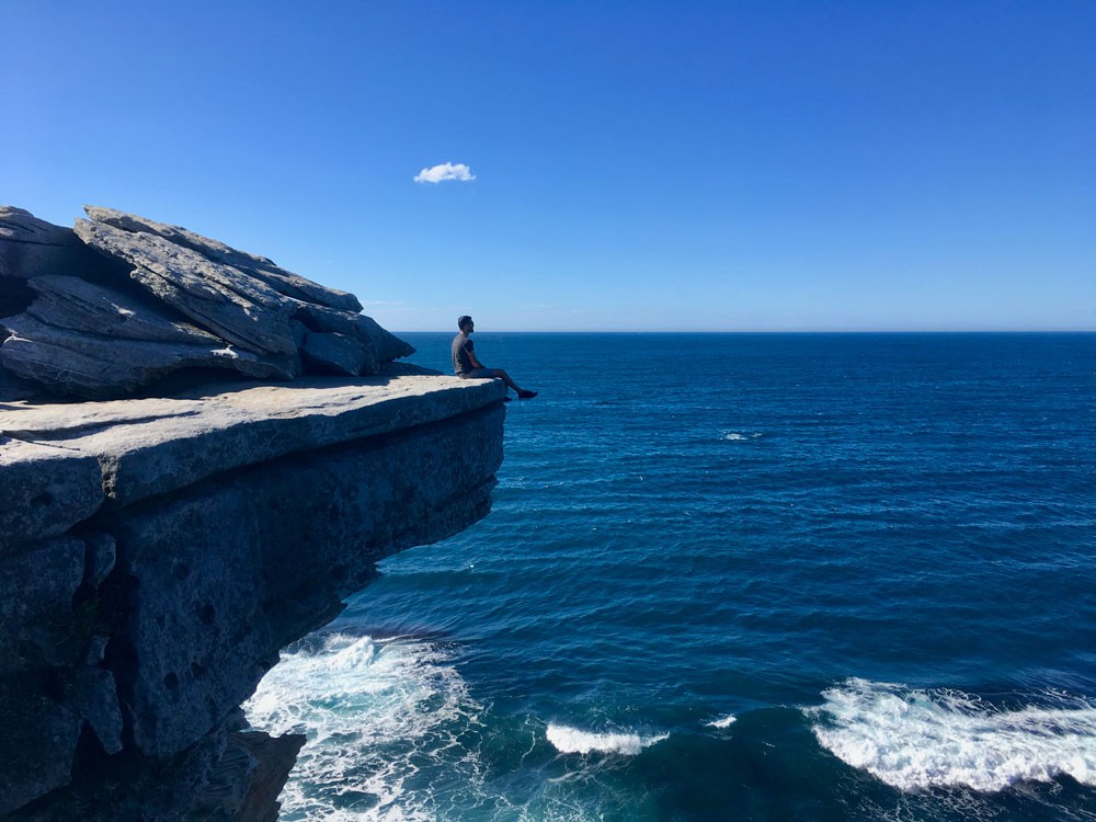 chico de espaldas sentado en una roca alta y debajo el mar 